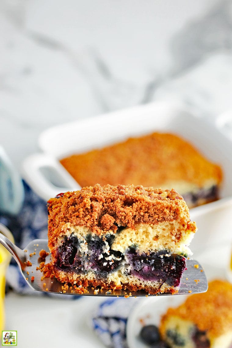 A slice of blueberry muffin coffee cake on a cake server with a baking dish and plate of cake in the background.