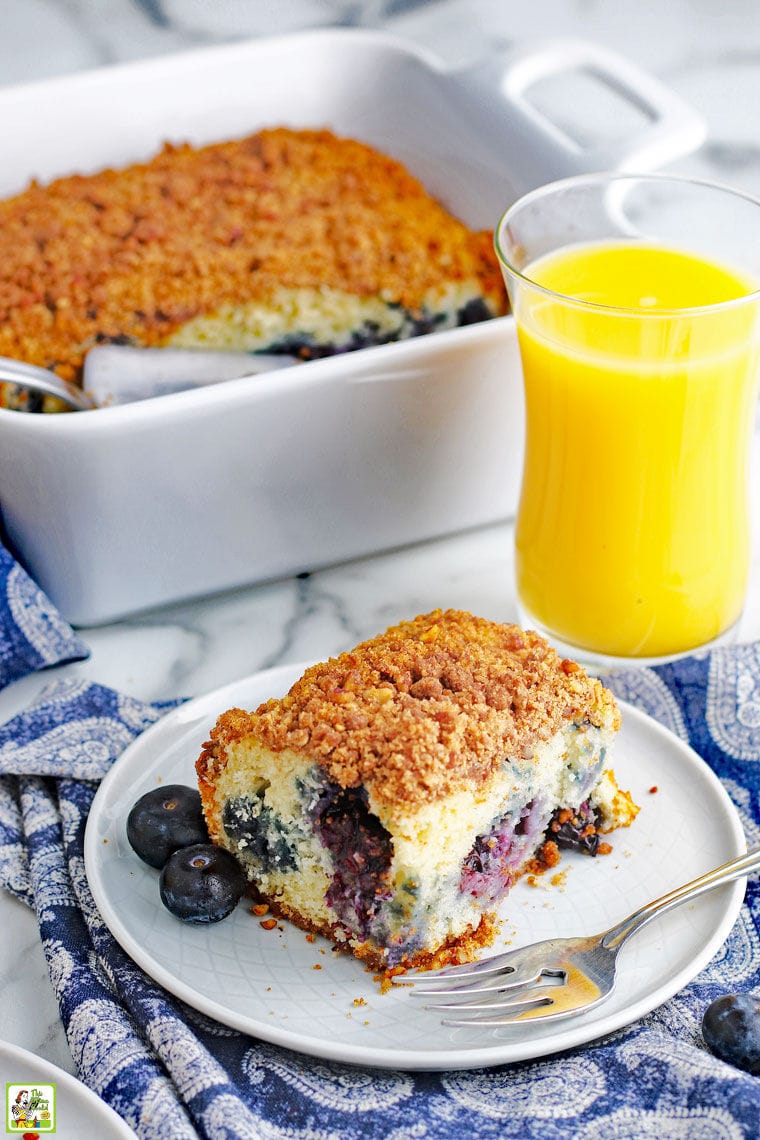 A piece of blueberry muffin coffee cake on a white plate with fork, a baking dish of coffee cake, and a glass of orange juice on a blue and white napkin.