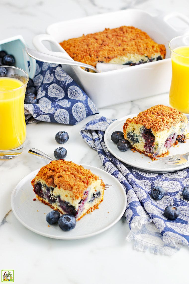 Pieces of blueberry muffin cake on white plates with fork, a baking dish of coffee cake, and glasses of orange juice on a blue and white napkins.