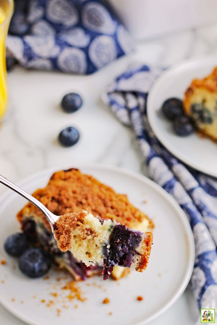 A forkful of coffee cake above a piece of blueberry muffin cake on a white plate with blue and white cloth napkins, blueberries, and a glass of juice.