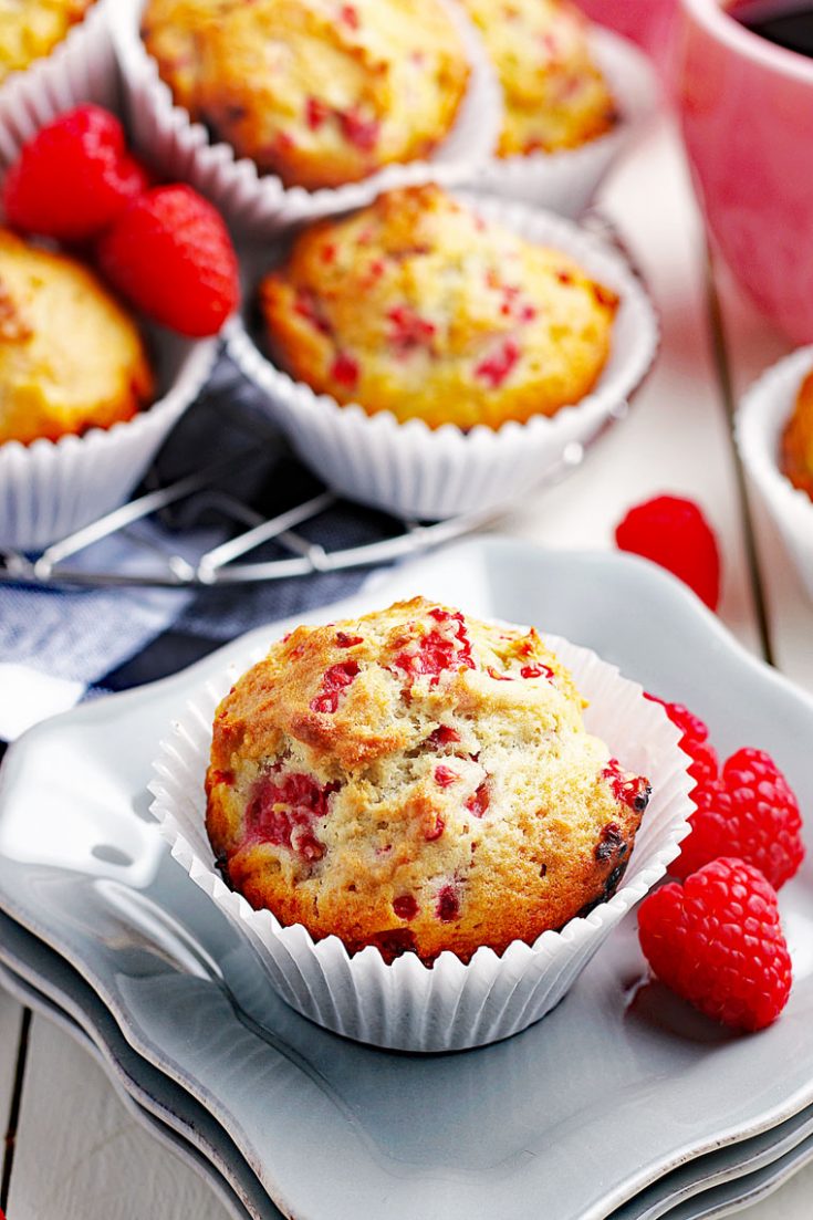 A raspberry muffin on white plates with fresh raspberries with more raspberry muffins in white cupcake liners in the background.