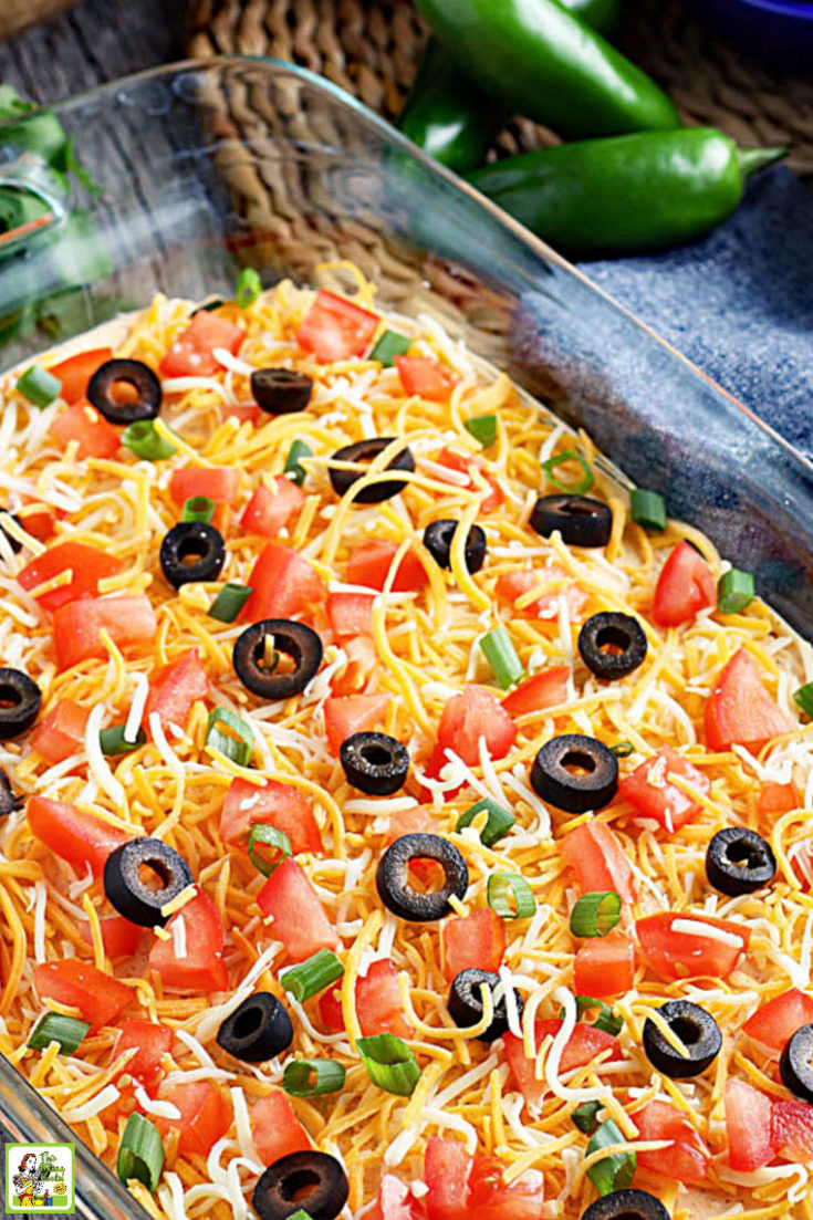 A casserole dish of layered taco dip with cheese, olives, tomatoes, and green onions.