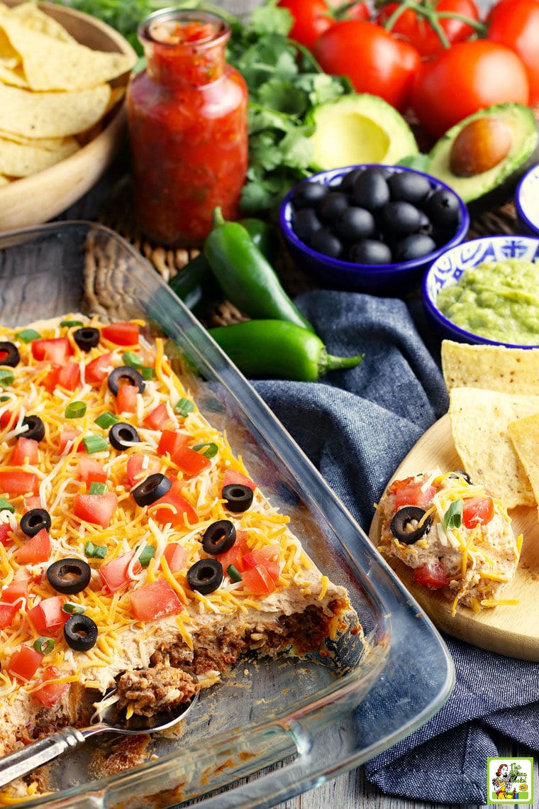 A glass casserole dish of layered party dip with layers of cheese, olives, tomatoes, green onions, sour cream, refried beans, and ground beef. With olivers, avocadoes, tortilla chips, chiles, and tomatoes in the background.