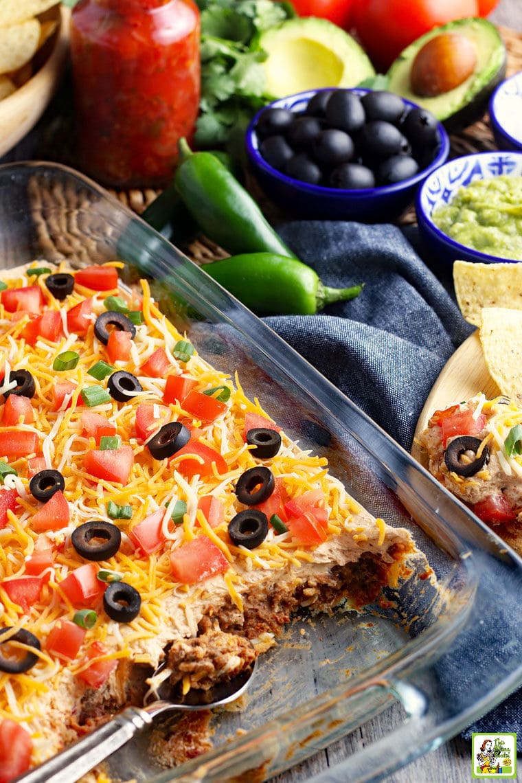 A casserole dish of with cheese, olives, tomatoes, and green onions with layers of ground meat and refried beans in a layered party dip.