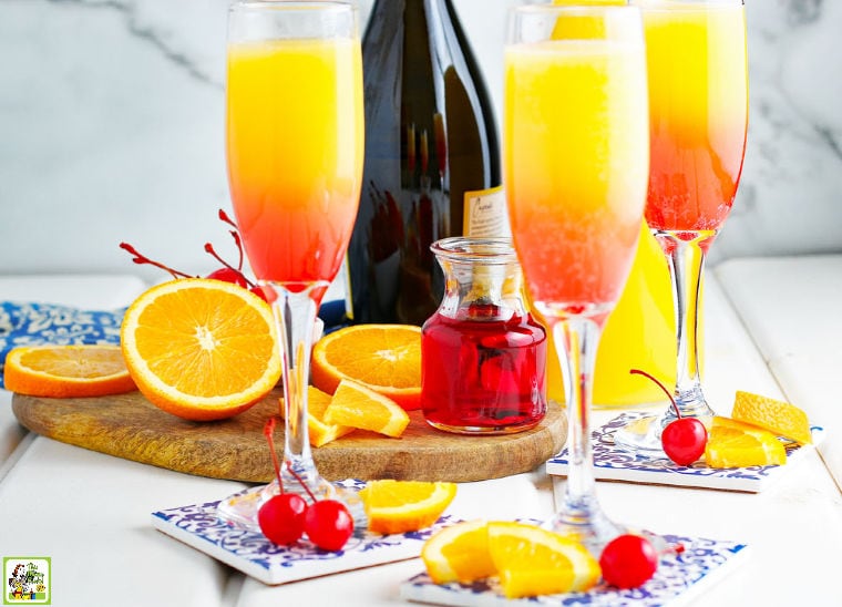 Glasses of prosecco mimosa with a champagne bottle, cherries, orange slices, coasters, and a wooden cutting board.