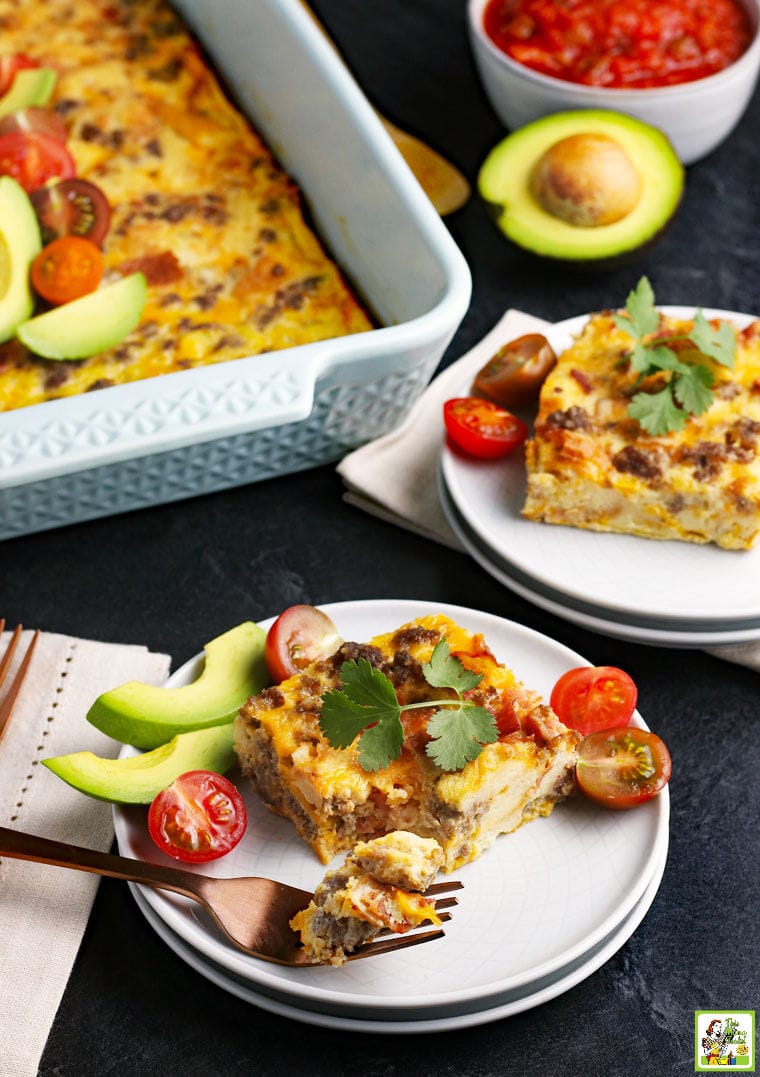 White plates a cheesy breakfast casserole with a large baking dish with tomatoes and avocados, forks and napkins.