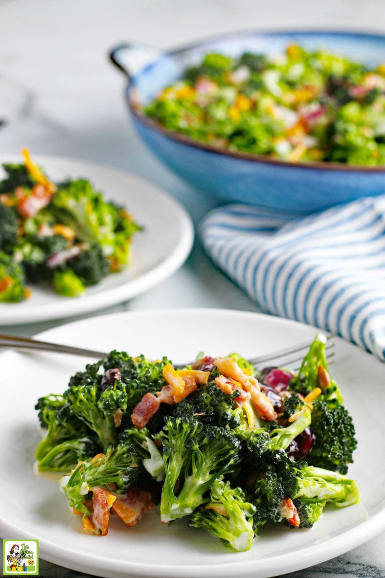 Two whites plates of Broccoli Salad with cheese, red onion, sunflower seeds, and cranberries. Blue and white napkins and a blue salad bowl in the background.