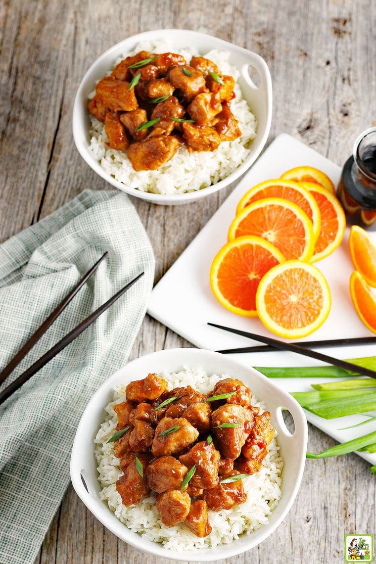Two bowls of Orange Chicken on white rice with slices of oranges, soy sauce, green onions, and chopsticks.