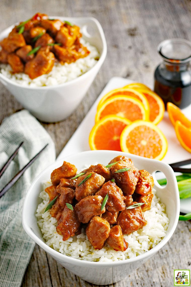 A bowl of Orange Chicken on white rice with slices of oranges, soy sauce, green onions, and chopsticks.