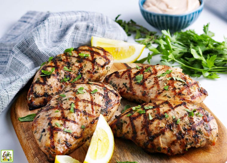 Four grilled marinated chicken breasts on a wooden cutting board with lemons and herbs. 