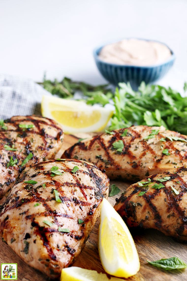 Grilled pieces of easy marinated chicken on a wooden cutting board, with lemon wedges, parsley, and a bowl of dipping sauce.