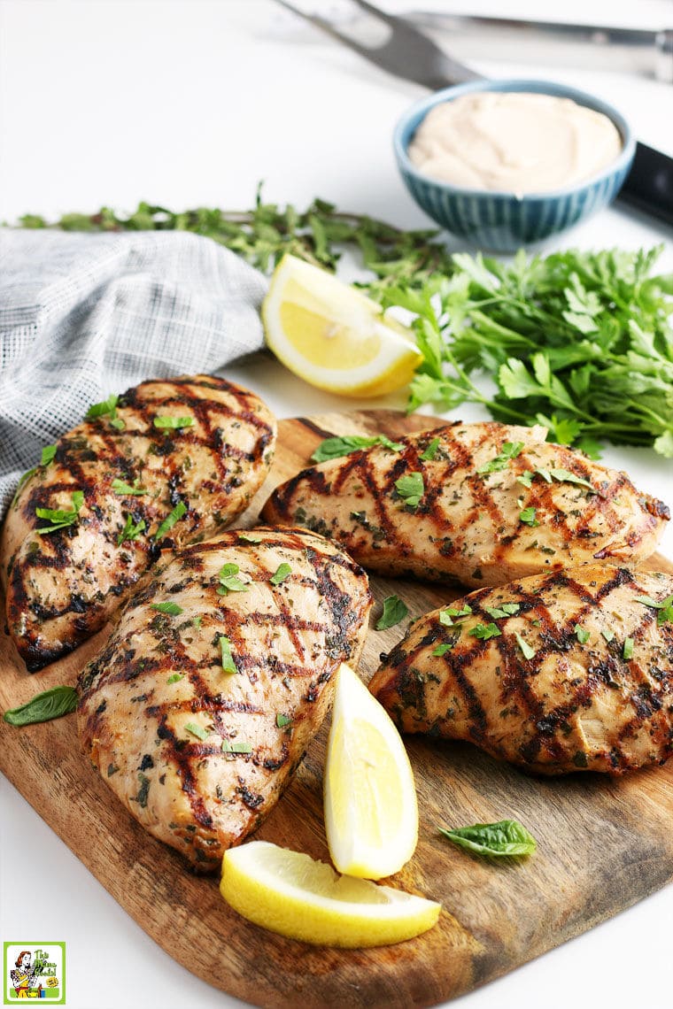 Grilled pieces of marinated chicken on a wooden cutting board, with lemon wedges, parsley, a bowl of dipping sauce, and grilling tools.