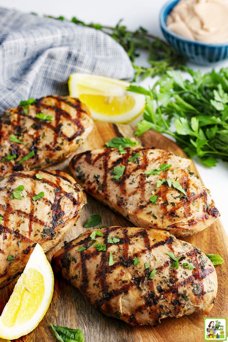 Four pieces of grilled easy marinated chicken on a wooden cutting board, with lemon wedges and parsley.