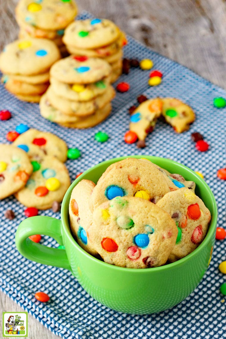 M&M Chocolate Chip Cookies in a green mug with stacks of M&M cookies in the background