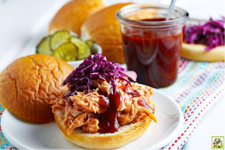 BBQ Chicken sandwich on a white plate with pickles with a jar of homemade BBQ, buns, purple cabbage in the background.