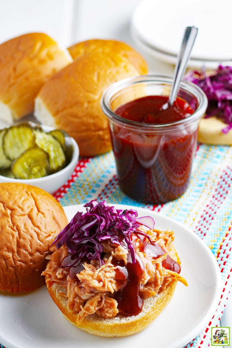 BBQ chicken sandwiches with red cabbage, pickles, BBQ sauce, and buns on a striped cloth
