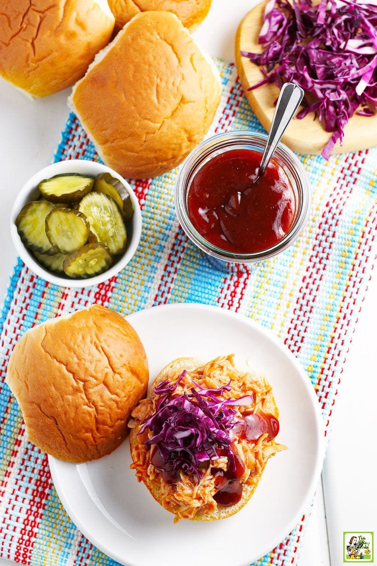 Overhead shot of BBQ chicken sandwiches, pickles, BBQ sauce, and buns on a striped tea towel