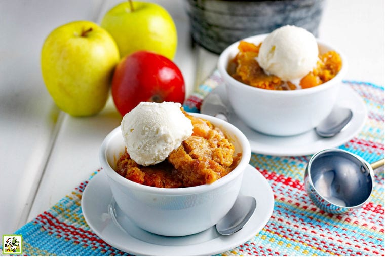 Saucers of Crockpot Apple Cobbler Recipe with scoops of vanilla ice cream on a multicolored woven placemat with spoons, apples, and an ice cream scoop.