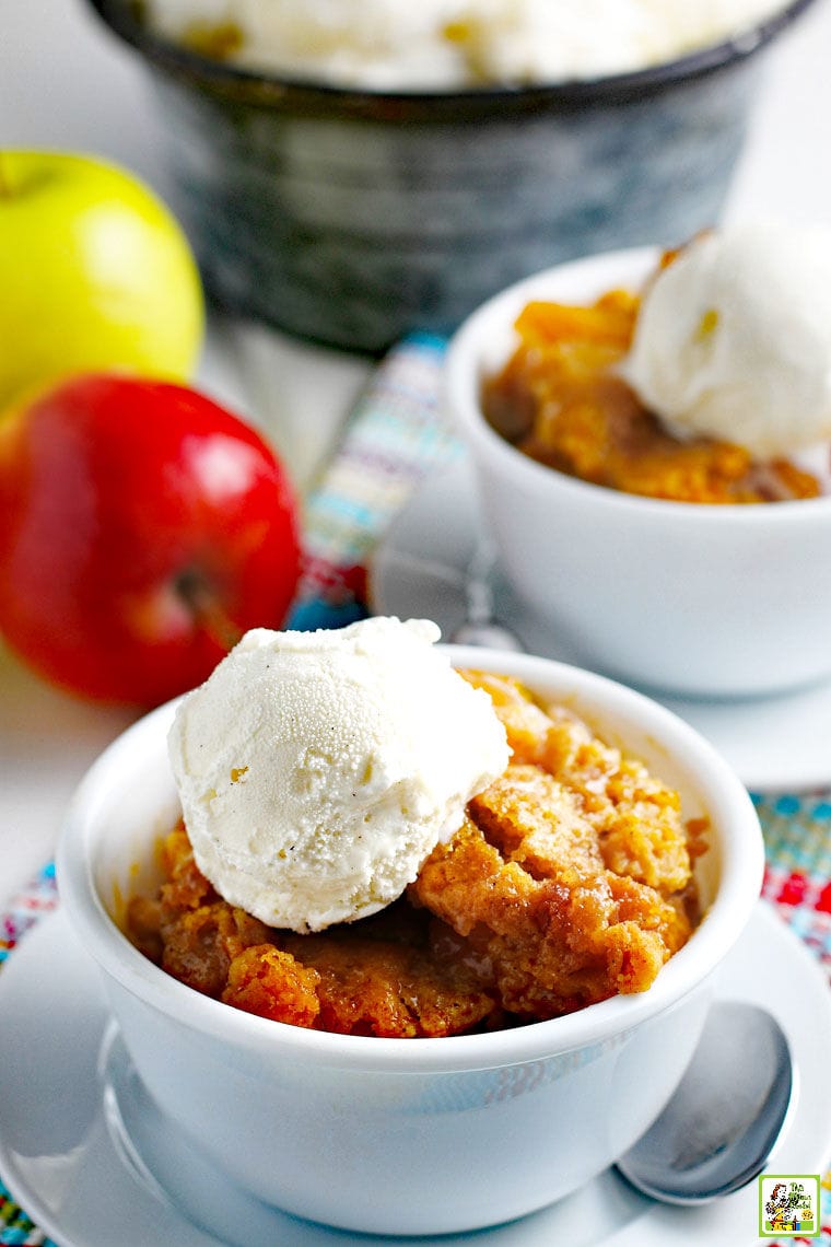 Apples and bowls of apple cobbler with a scoop of vanilla ice cream with a slow cooker in the background.