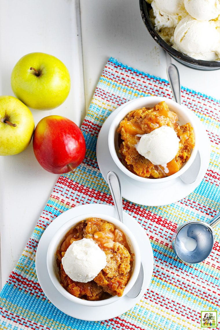 Overhead shot of two bowls of cobbler with apples, an ice cream scoop, on a brightly colored placemat.