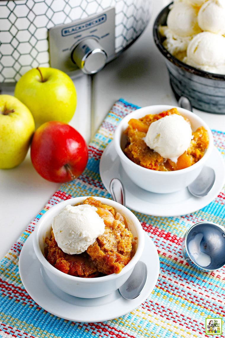 Apples, bowls of cobbler with ice cream, an ice cream scoop, on a brightly colored placemat with a slow cooker in the background.