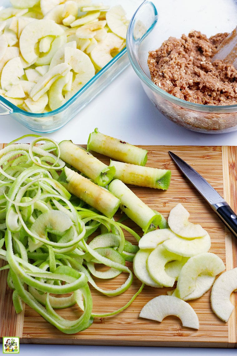 Ingredients for apple crisp - cored, peeled and sliced apples on a wooden cutting board with a paring knife, apple crisp topping, and sliced apples in a glass baking pan.