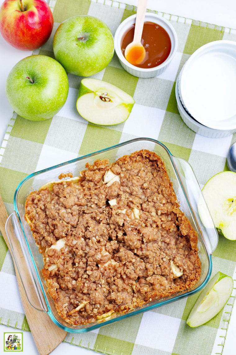 A dish of Apple Crisp with bowls, spoons and apples.