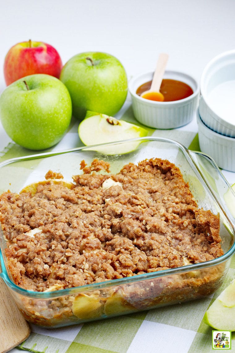 A platter of warm apple crisp with apples, apple slices, a bowl of caramel sauce, and white bowls on a green and white napkin.