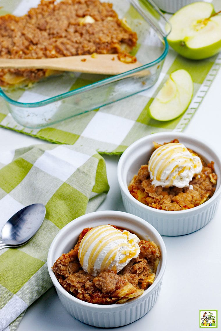 Bowls of apple crisp with scoops of vanilla ice cream, a pan of apple crisp with a wooden serving spoon, green and white napkins and sliced apples.