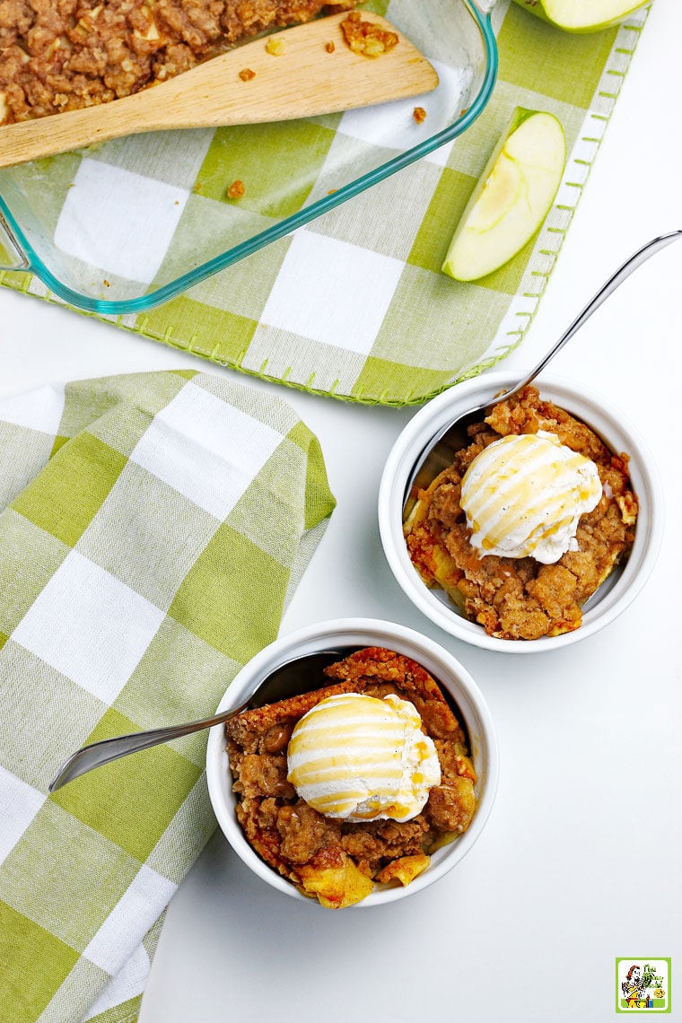 Overhead view of two bowls of fruit crisp with ice cream on green and white napkin with a glass baking pan and sliced apples.