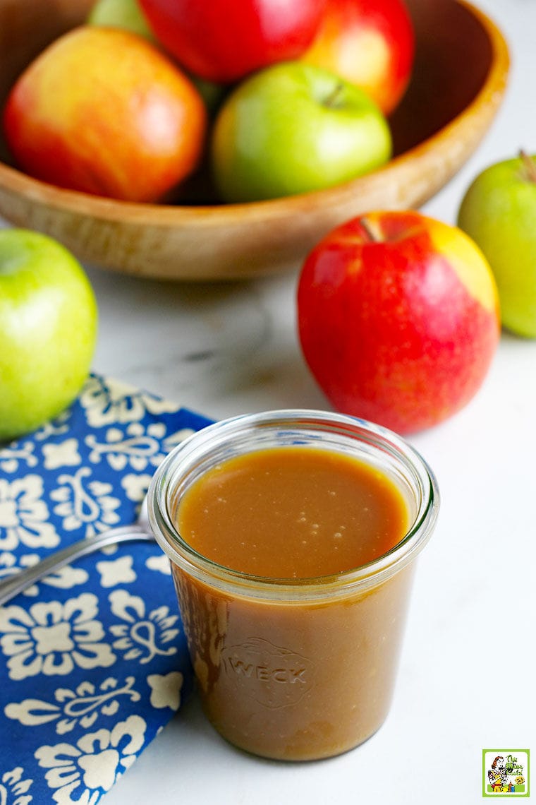 A glass jar of homemade vegan caramel sauce, a bowl of apples, a spoon, and a blue and white napkin.