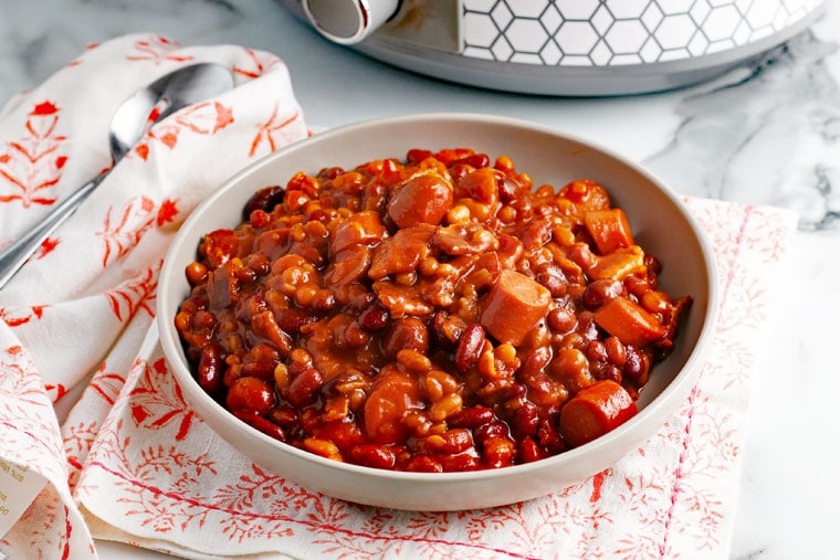 A bowl of Crock-Pot Baked Beans on a napkin with a spoon with a slow cooker in the background.