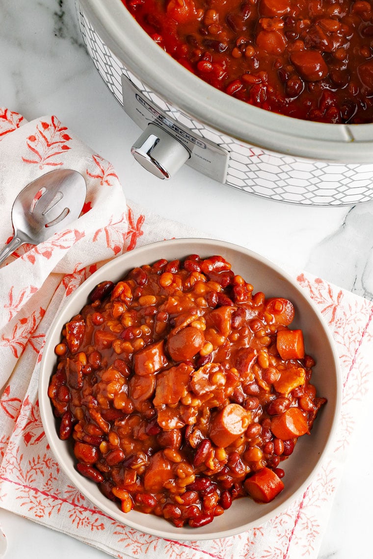 Overhead shot of a bowl of Crock-Pot Baked Beans with orange and white napkins, spoon, and a slow cooker filled with baked beans.