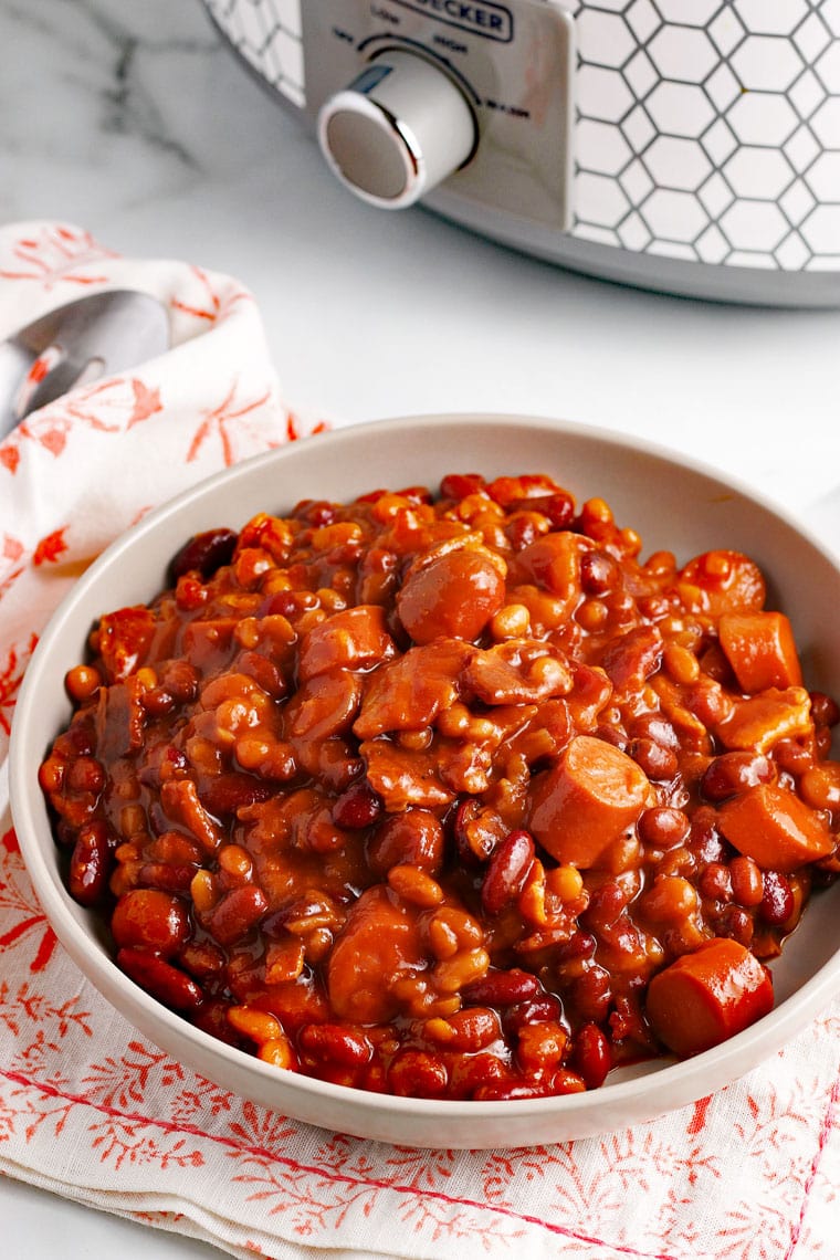 Bowl of slow cooker baked beans with hot dogs on a white tabletop with orange and white floral napkins with silver spoon and white and sliver slow cooker.