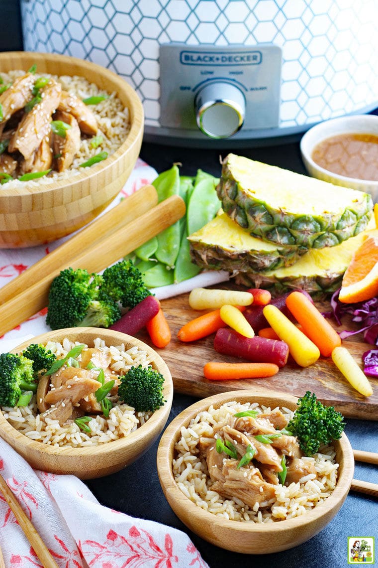 A slow cooker, serving bowls of teriyaki chicken on rice with broccoli, wooden serving utensils, wooden cutting board, fruits and vegetables, on colorful napkins. 