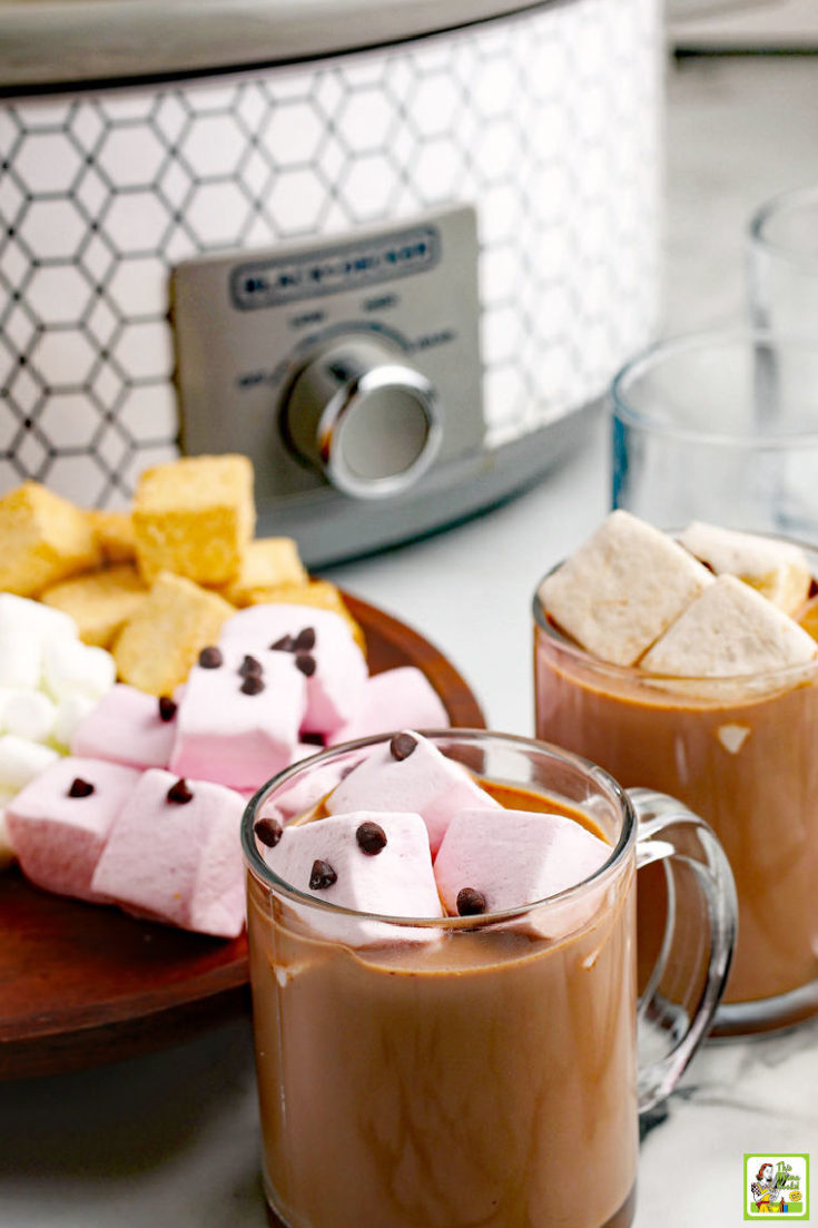 Mugs of hot chocolate with marshmallows in glass mugs with a slow cooker in the background.