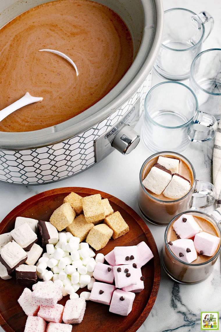 Overhead shot of a crock pot of hot chocolate with a ladel, a wooden cutting board covered in colorful marshmallows, and glass mugs of hot chocolate and empty mugs on a marble countertop.
