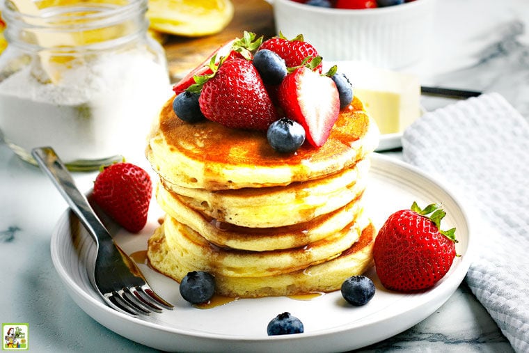 A plate of fluffy pancakes with berries and syrup, a fork, napkin, and a jar of dry pancake mix in the background.