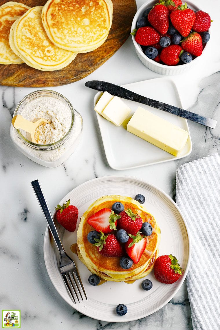 Overhead view of a stack of pancakes with fruit and syrup on a white plate, a glass jar of pancake mix, a stick of butter with a knife on a white ceramic dish, a wooden cutting board with pancakes, and a bowl of fruit.