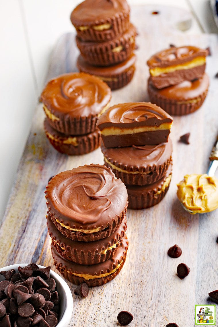 Stacks of hand made peanut butter cups with a spoon of peanut butter and a bowl of chocolate chips on a wooden board.