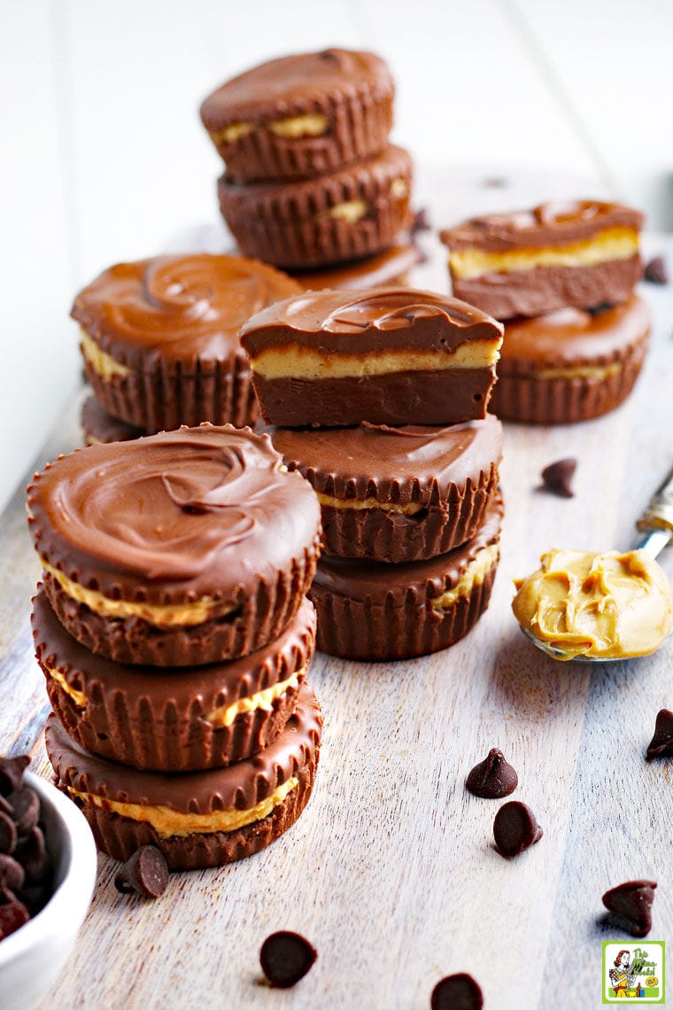 A wooden cutting board with stacks of peanut butter cups, bowl of chocolate chips, and a spoon of creamy peanut butter. 