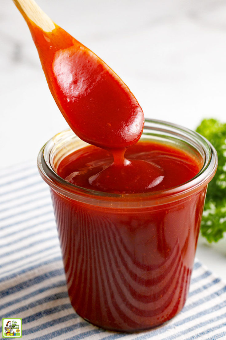 Wooden spoon dipping into glass jar of sugar free barbecue sauce with parsley in the background.