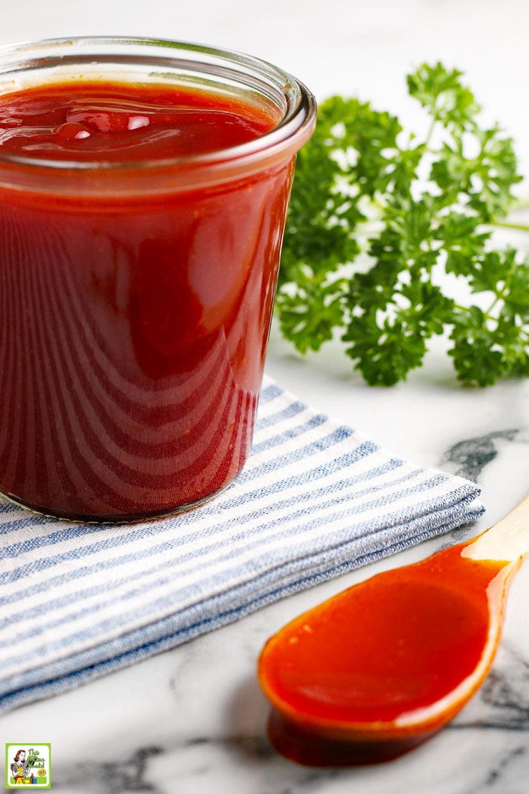 Glass jar of keto barbecue sauce on a blue and white striped napkin and a wooden spoon covered in BBQ sauce.