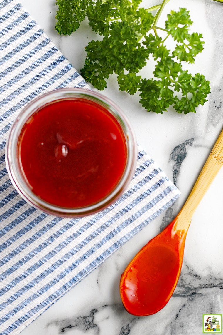 Overhead shot of a glass jar of homemade barbeque sauce, wooden spoon covered in sauce, blue and white striped napkin, and a sprig of fresh parsley.