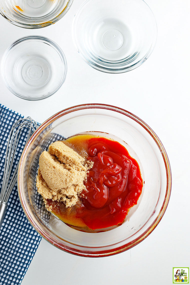 Making low carb BBQ sauce in a glass bowl with whisk, small glass bowls of ingredients on a blue and white cloth napkin.