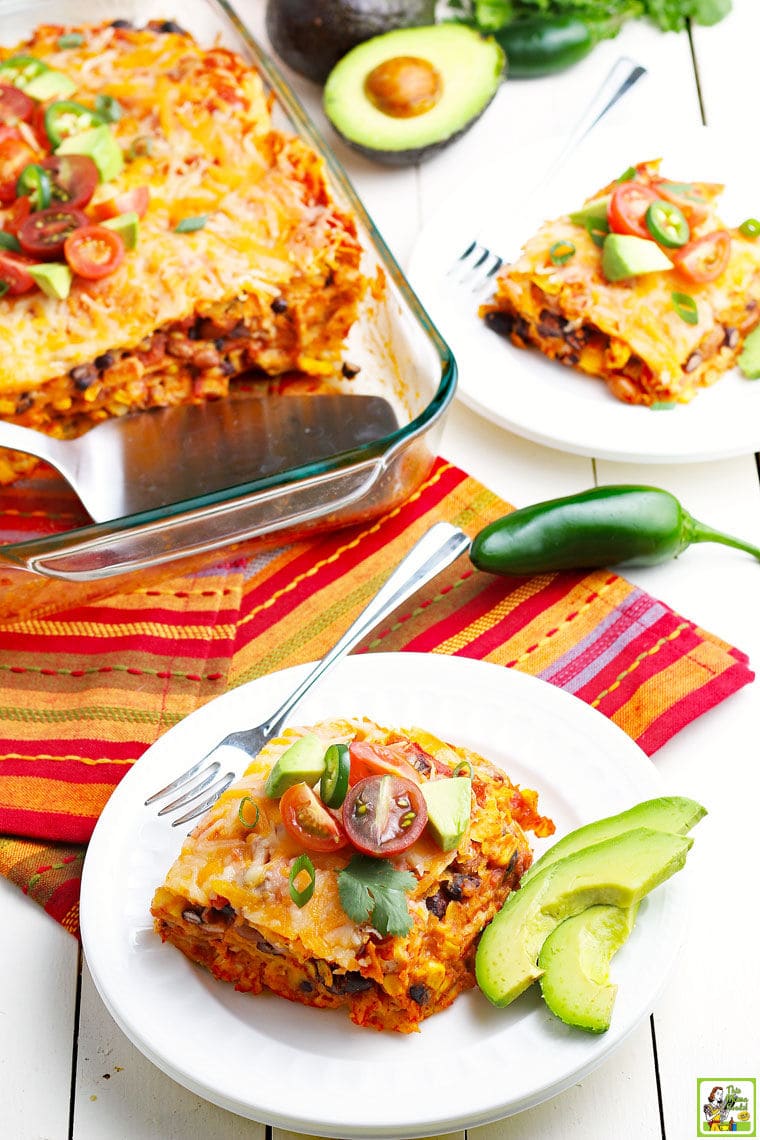A glass casserole dish of chicken enchilada with white plates of slices of chicken enchilada with chili peppers, avocado, and tomatoes on festive napkins.