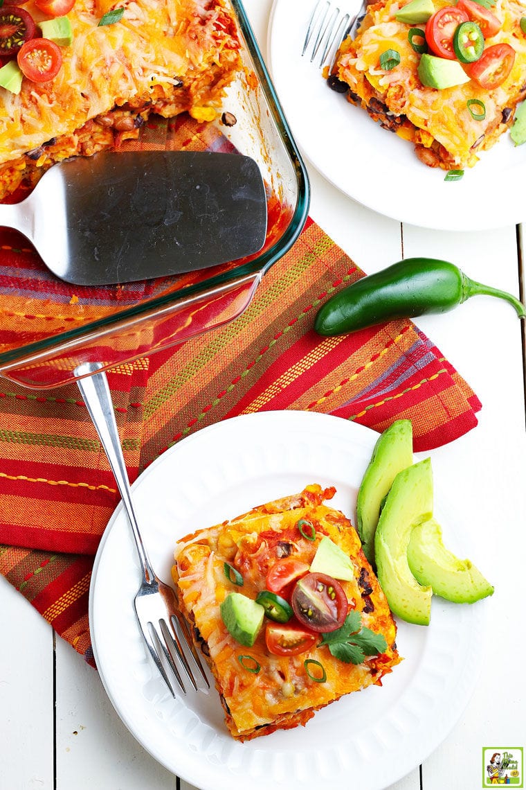 A square slice of enchilada casserole on a white plate with slices of avocados and cherry tomatoes with a glass casserole dish of enchiladas with a silver serving spoon placed on a colorful napkin.