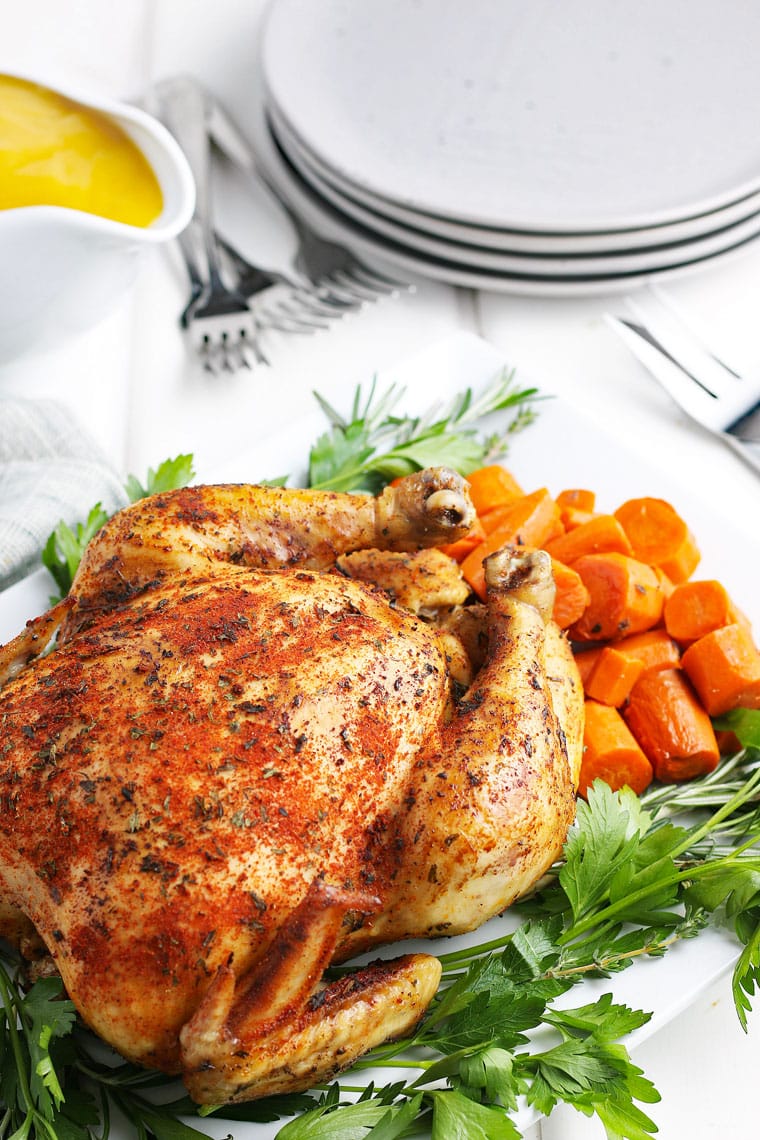 A crockpot whole chicken on a platter with carrots, gravy, plates, and forks.