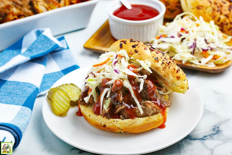 An Instant Pot Pulled Pork sandwich on a plate with pickle, napkin and more in the background with a ramekin with BBQ sauce.