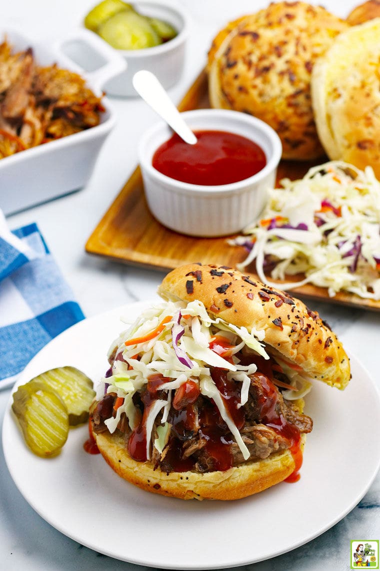 A barbecue pork sandwich with coleslaw on a white plate with pickles with a blue and white napkin with sandwich fixings and a platter of shredded pork.